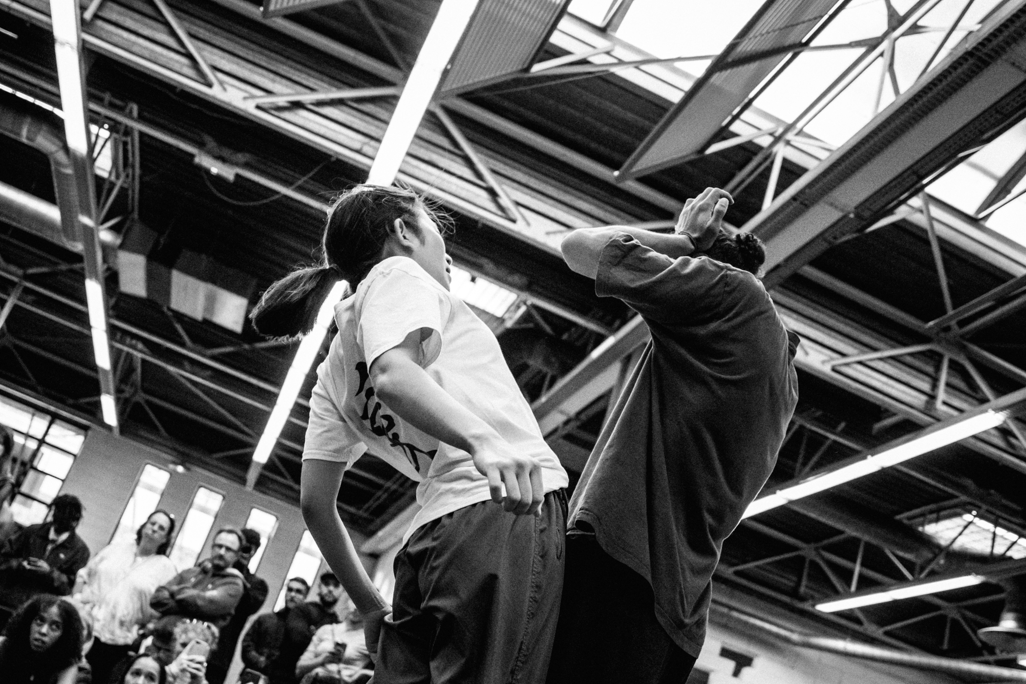 Deux danseurs s'affrontent dans une battle, entourés d'un public attentif sous une grande verrière industrielle
